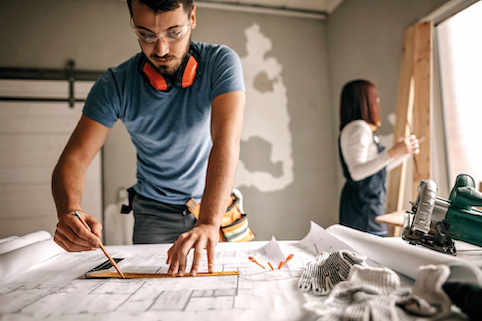 Man reviewing construction plans while woman in background is working on framing construction project.