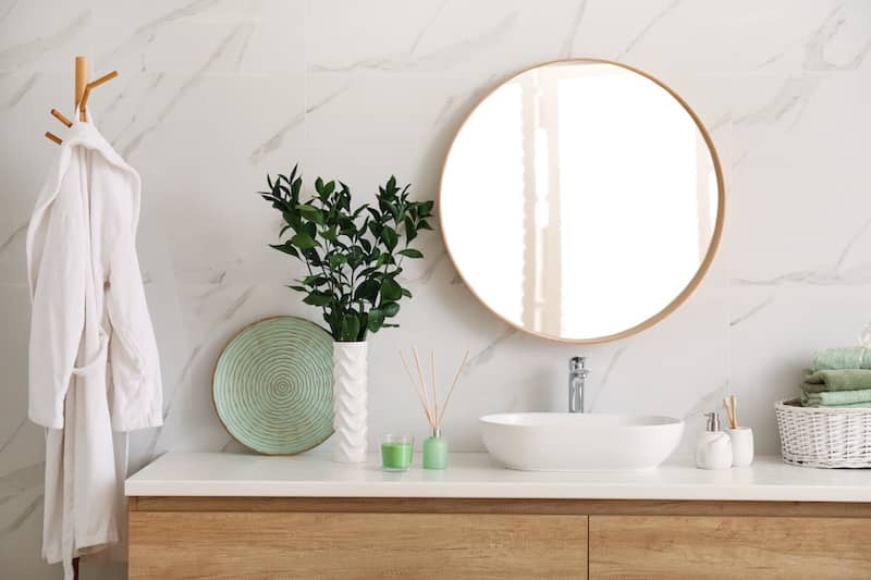 Bathroom vanity with circular mirror, a candle, and fresh green sprigs in white vase.