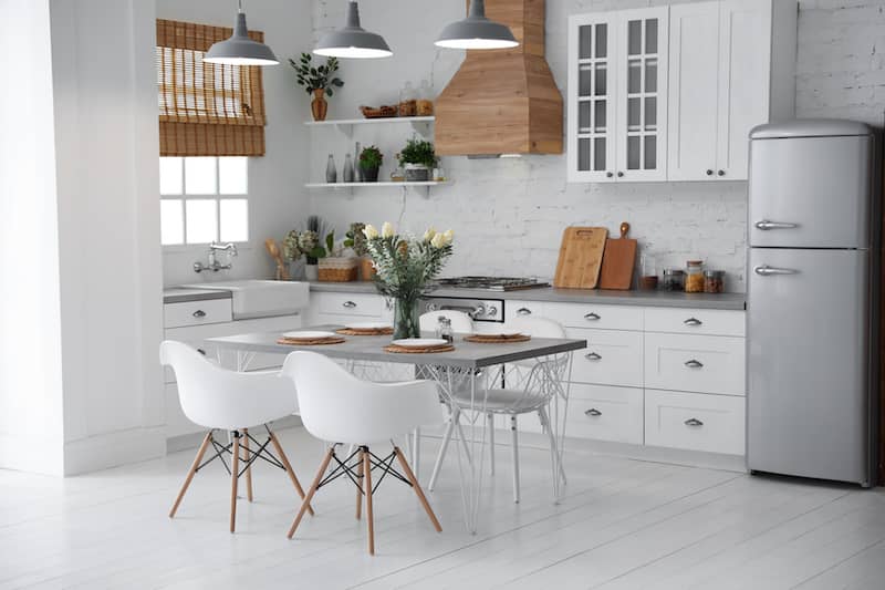 White kitchen with bamboo shades pulled up on window, white midcentury modern chairs at kitchen table and small open shelves with potted plants. A vase of fresh flowers is on the kitchen table.