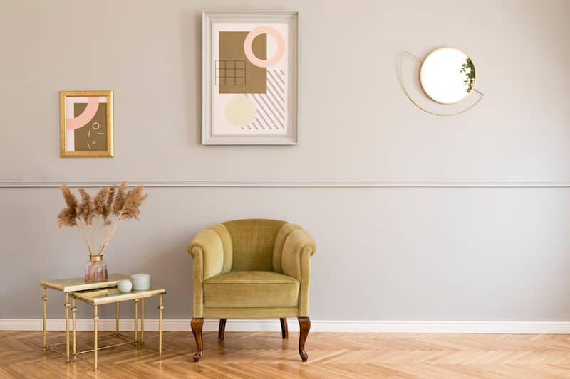 Stylish wall art and mirror on grey wall over small velvet chair next to gold nesting tables with natural dried elements in vase.