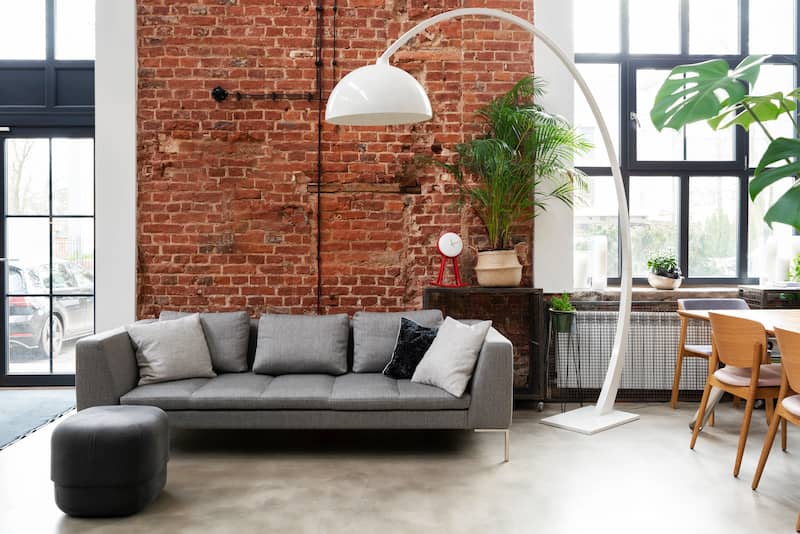 Loft style interior with large windows with exposed brick wall, grey couch, and large midcentury style floor lamp arching over the couch.
