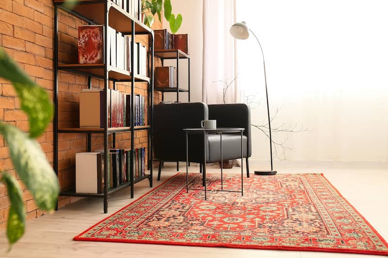 Reading nook in living room featuring antique red rug, black leather chair, vintage floor lamp, bookshelves and a large houseplant near a window with light filtering through white sheer curtains.