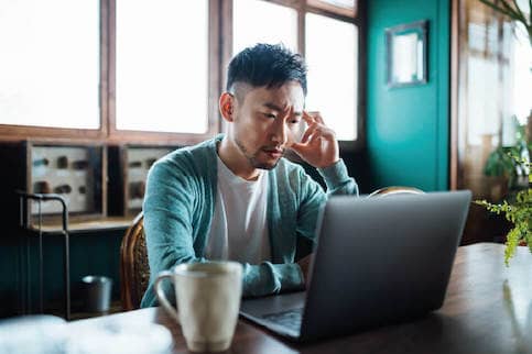 Distressed looking Asian man sitting at a table using a laptop.