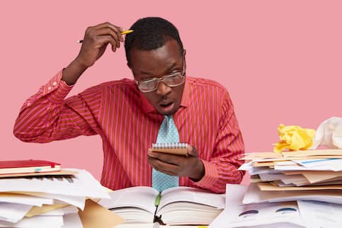 African American man scratching his head with a pencil while looking at a notepad and surrounded with books.