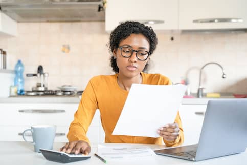 Young African American woman in a yellow shirt budgeting at home with a calculator and paper.