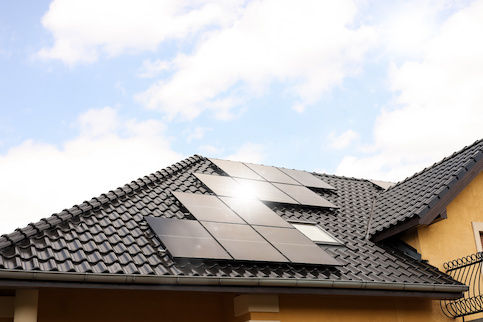 Black gabled roof with solar panels with blue sky with clouds and sun peeking through in background.