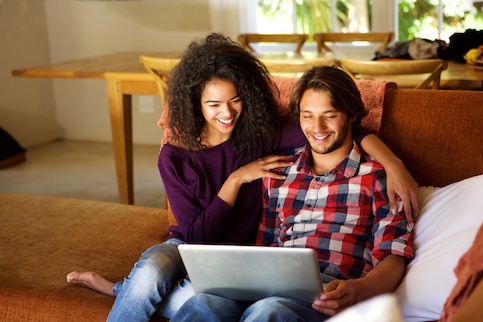 A couple looking at information on a laptop on their couch, smiling at the information on the screen.