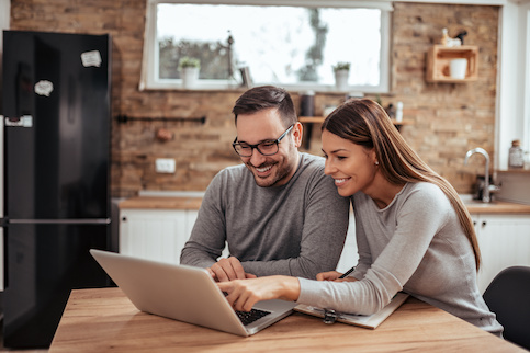 Smiling couple looking at laptop on kitchen table together with paperwork nearby.