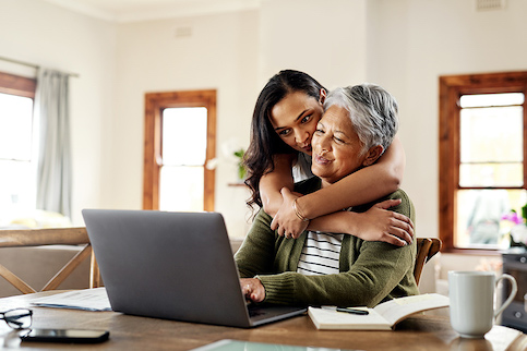 Adult daughter hugging mother at laptop, smiling, at dining room table in home.