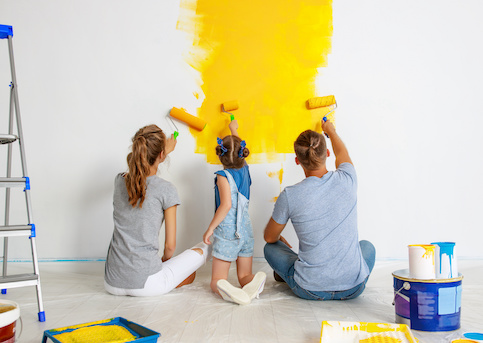 Mother, young daughter and father painting white wall yellow with paint cans and ladder nearby.