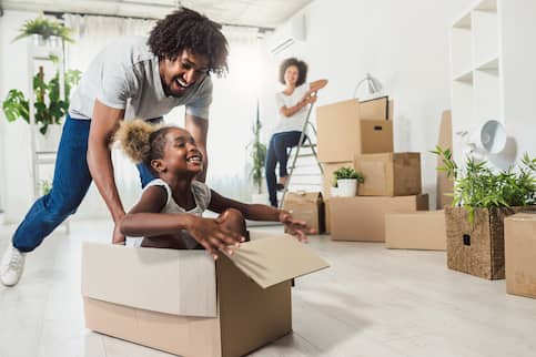 Smiling father pushing laughing daughter amongst other moving boxes in new home.