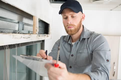 Man in cap and gray shirt inspecting home with a clipboard.