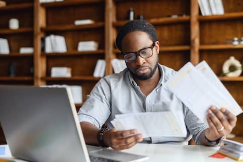 Young African American man looking through papers in front of laptop at desk.