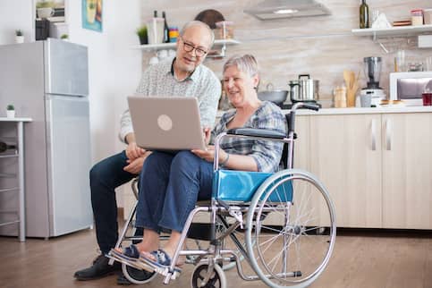 Older couple in their kitchen with one in a wheelchair and using a laptop.