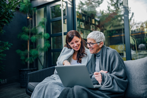 Older mother with a silver pixie cut looking at laptop with daughter on patio couch.