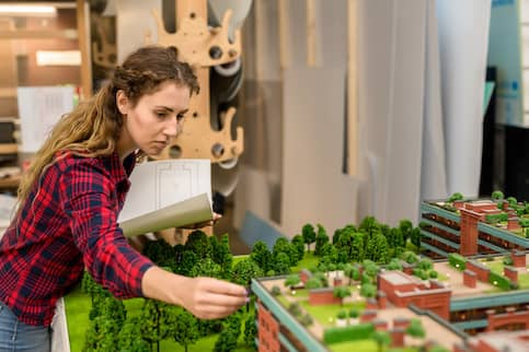 Young woman with blueprint looking at one of modern architectural constructions on city layout model.