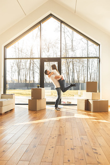 Couple in new home in front of picture window embracing with moving boxes nearby.