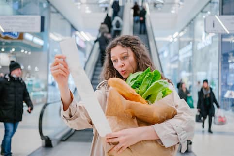 Woman in a store holding a receipt and grocery bag while looking shocked.
