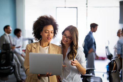 Two female realtors looking at laptop standing up in office setting.