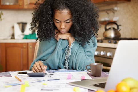 Young African American woman budgeting with calculator and laptop at home in kitchen.