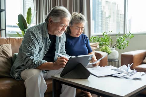 Senior couple going over financial paperwork together while sitting on couch in living room with houseplants.