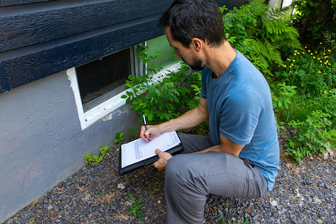 Contractor or inspector inspecting home with list of items. Crouching down to examine basement windows.