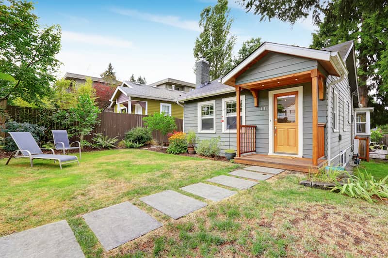 Gray, one-story house with wooden porch and door leading out to yard with lawn chairs and grey stepping stones.