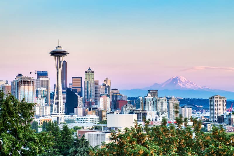 View of Seattle skyline with large trees in foreground and mountain range in background.