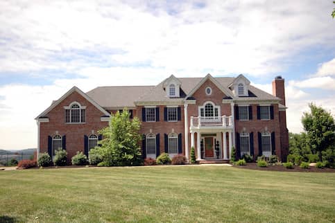 Large, two-story brick McMansion with ornate balcony over front door and large white columns. 