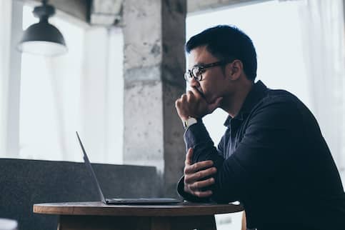 Asian man looking pensively at his laptop with his hand over his mouth.