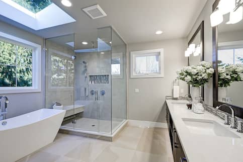 Light gray bathroom with large white tub, separate glass shower, two windows and large tile floor.
