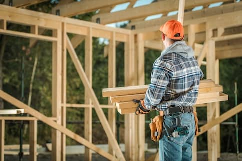 Construction worker in hard hat holding wooden beams looking at frame of new house.