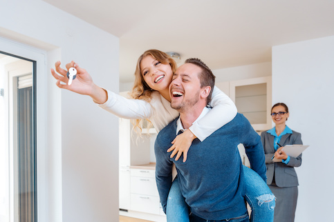 Man carrying woman holding up house keys and celebrating closing on a house as real estate agent in background smiling.