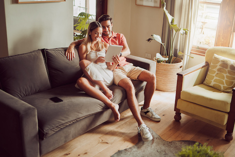 Young couple sitting on couch looking at tablet, smiling.