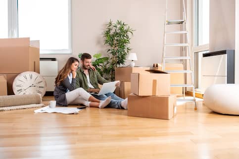 Young couple sitting on floor with computer on lap and moving boxes all around them.