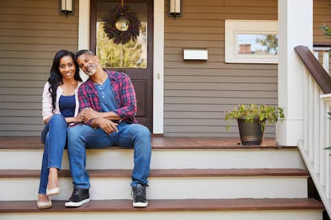 Middle aged couple sitting on wood porch steps, holding hands and smiling.