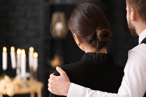 Young man and woman dressed in black and with backs to camera, comforting each other in front of lit candles.