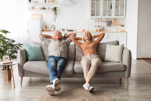 Senior couple relaxing on grey sofa and smiling with arms behind their heads.
