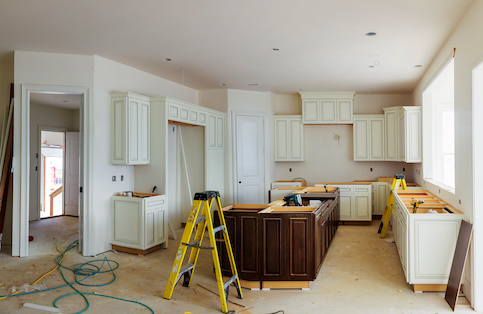 : Kitchen in the process of remodel with bare floors, yellow ladder, wires and cabinets painted white and brown with no countertops.
