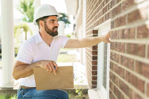 Man wearing white hard hat and holding clipboard looking at exterior of home.