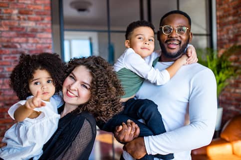 Happy multiethnic family of four standing outside red brick home.