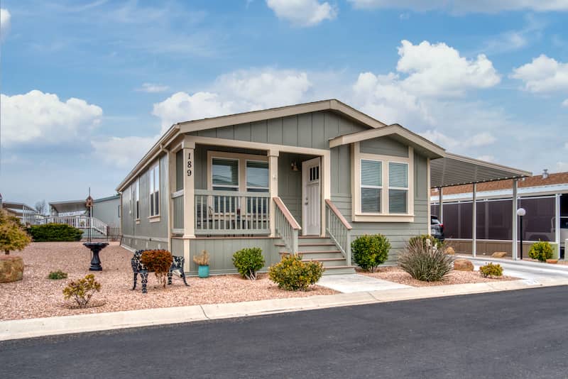 Sage green manufactured home with tan trim sitting on lot with wood chip landscaping and bench.