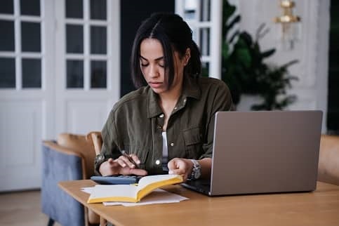 Young woman sitting at table with laptop open, using calculator and reviewing notebook.