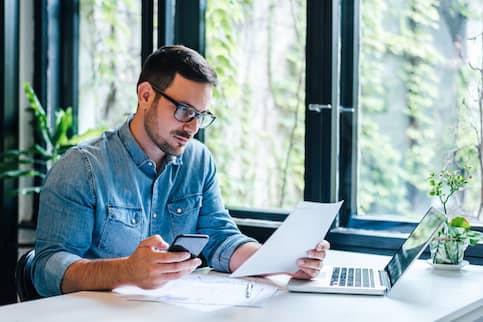 Young man wearing glasses, holding cell phone and looking at paper documents while sitting at desk with open laptop.