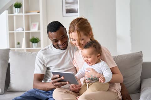 Small multiracial family with baby sitting on the couch together and playing with a phone or tablet.