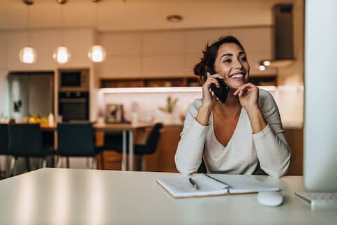 Young woman with open notebook, sitting in front of computer in kitchen talking on phone 