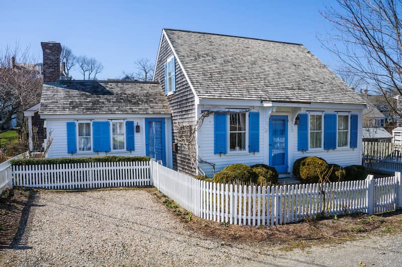 A low white picket fence surrounds the small lot of a blue shuttered cottage at the tip of Cape Cod.