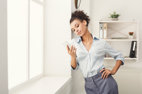 Woman leaning against wall near window checking phone with hand on hip.