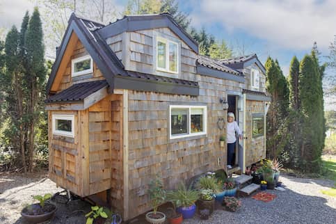 Older woman in doorway of charming tiny home with large potted plants out front.