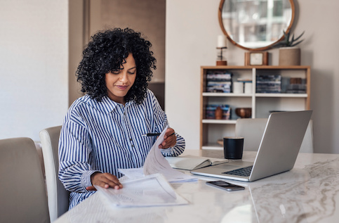 Woman reviewing paperwork with open laptop at home office desk.